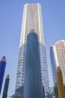 A perspective view of tall, colorful pencil sculptures standing in front of high-rise buildings. The sky is clear and blue, providing a clean background that contrasts with the vibrant colors of the pencils and the buildings' sleek architecture.