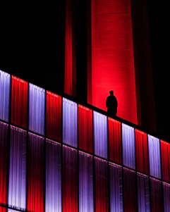 A sleek, shadowed figure wearing matte black streetwear with neon red accents, standing against a backdrop of a dimly lit cityscape at night.
