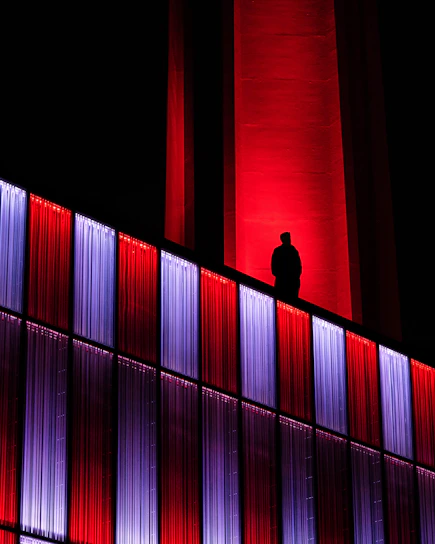 A sleek, shadowed figure wearing matte black streetwear with neon red accents, standing against a backdrop of a dimly lit cityscape at night.