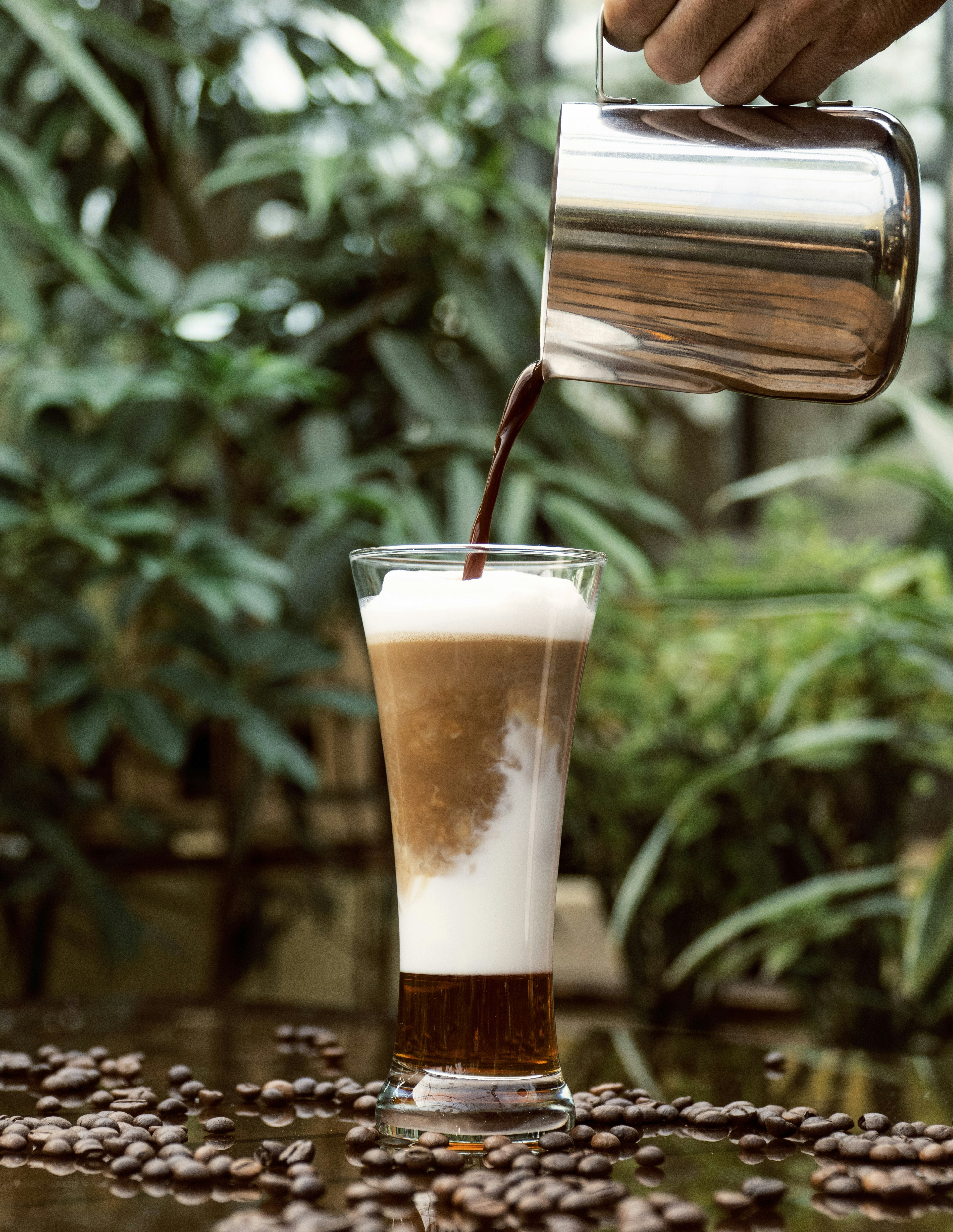 A creamy coffee beverage is being poured into a tall glass, surrounded by scattered coffee beans on a reflective surface. Green plants are blurred in the background, adding a natural atmosphere.