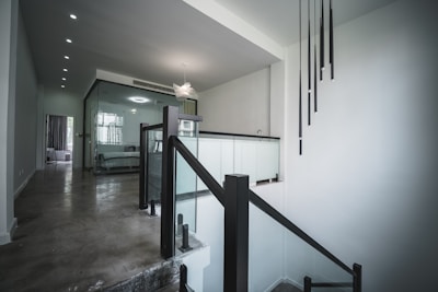 A modern interior space featuring a staircase with black railings and glass panels. A minimalist lighting fixture hangs from the ceiling. The walls are painted white, and the floor is a polished concrete. In the background, there is a room with a plush bed visible through a glass partition.