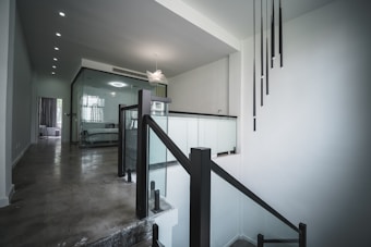 A modern interior space featuring a staircase with black railings and glass panels. A minimalist lighting fixture hangs from the ceiling. The walls are painted white, and the floor is a polished concrete. In the background, there is a room with a plush bed visible through a glass partition.
