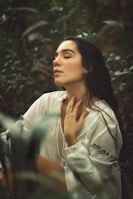 A person with long dark hair is seated in a lush, green forest environment, wearing a white, flowing blouse. They appear serene and relaxed, with eyes closed and one hand gently touching the neck.