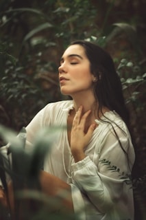 A person with long dark hair is seated in a lush, green forest environment, wearing a white, flowing blouse. They appear serene and relaxed, with eyes closed and one hand gently touching the neck.