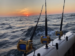 Three fishing rods with reels are mounted on the side of a boat, set against a backdrop of a sunset over the ocean. The water appears choppy with visible waves, and the sky transitions from a soft orange near the sun to a darker hue at the top.