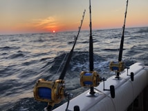 Three fishing rods with reels are mounted on the side of a boat, set against a backdrop of a sunset over the ocean. The water appears choppy with visible waves, and the sky transitions from a soft orange near the sun to a darker hue at the top.