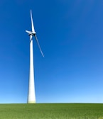 A large wind turbine stands prominently against a clear blue sky, situated on a lush green field. Its blades are positioned to capture wind energy efficiently.