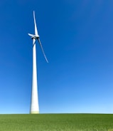 A large wind turbine stands prominently against a clear blue sky, situated on a lush green field. Its blades are positioned to capture wind energy efficiently.