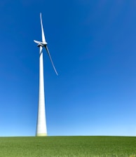 A large wind turbine stands prominently against a clear blue sky, situated on a lush green field. Its blades are positioned to capture wind energy efficiently.