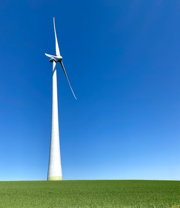 A large wind turbine stands prominently against a clear blue sky, situated on a lush green field. Its blades are positioned to capture wind energy efficiently.