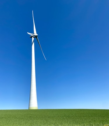 A large wind turbine stands prominently against a clear blue sky, situated on a lush green field. Its blades are positioned to capture wind energy efficiently.