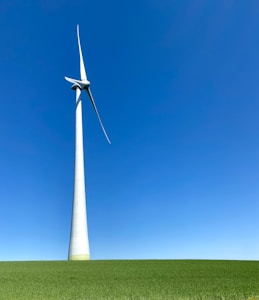 A large wind turbine stands prominently against a clear blue sky, situated on a lush green field. Its blades are positioned to capture wind energy efficiently.