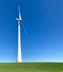 A large wind turbine stands prominently against a clear blue sky, situated on a lush green field. Its blades are positioned to capture wind energy efficiently.