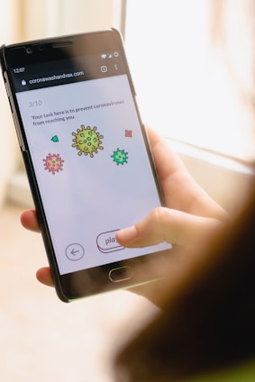 A hand holding a smartphone displaying a webpage with cartoon images of coronavirus particles. The text on the screen instructs the user to prevent the viruses from reaching them, indicating a game or interactive task. The interface includes a play button and navigation arrows.