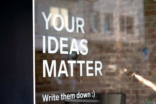 Text on a window with the words 'YOUR IDEAS MATTER' and 'Write them down :)'. The background shows a blurred brick wall.