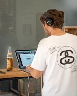 A man wearing headphones and a white graphic T-shirt is working on a laptop at a wooden table. A bottle of iced tea and a smartphone are placed nearby. The laptop screen displays text and images from a document. The setting appears to be a modern workspace with a reflective glass wall and some framed artwork in the background.
