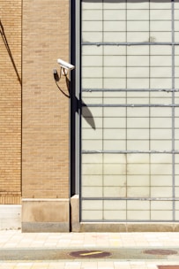 A security camera is mounted on the corner of a brick wall, casting a strong shadow. The brick wall is adjacent to a glass block wall, creating a stark contrast between the two surfaces. The paving in front of the walls includes tiles of varying shades.