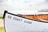 A wooden lifeboat with the words 'US Coast Guard' painted on its side rests on dry land. It is positioned at an angle that captures part of its interior, which is a natural wood color. In the background, there is a bright, sparkling view of the sea under a cloudy sky.