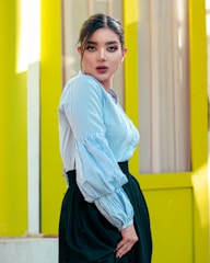 A person with dark hair styled in an updo wearing a blue and white striped blouse and a black skirt, standing confidently with a backdrop of vertical yellow panels that create a vivid, modern aesthetic.