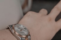 A close-up view of a luxury wristwatch being worn on a person's wrist. The watch has a metallic band and a round face, featuring a silver and rose gold color scheme. The watch face displays both the time and a date indicator.