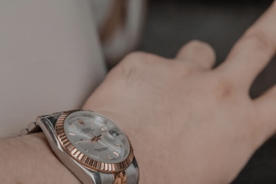 A close-up view of a luxury wristwatch being worn on a person's wrist. The watch has a metallic band and a round face, featuring a silver and rose gold color scheme. The watch face displays both the time and a date indicator.