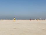A group of people wearing colorful sports uniforms are playing cricket on a wide, open sandy ground. The sky is clear with a subtle gradient from blue to grey on the horizon. In the distance, there are structures that suggest a cityscape.