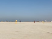 A group of people wearing colorful sports uniforms are playing cricket on a wide, open sandy ground. The sky is clear with a subtle gradient from blue to grey on the horizon. In the distance, there are structures that suggest a cityscape.