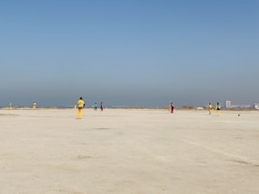 A group of people wearing colorful sports uniforms are playing cricket on a wide, open sandy ground. The sky is clear with a subtle gradient from blue to grey on the horizon. In the distance, there are structures that suggest a cityscape.