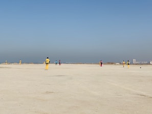 A group of people wearing colorful sports uniforms are playing cricket on a wide, open sandy ground. The sky is clear with a subtle gradient from blue to grey on the horizon. In the distance, there are structures that suggest a cityscape.