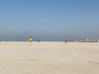 A group of people wearing colorful sports uniforms are playing cricket on a wide, open sandy ground. The sky is clear with a subtle gradient from blue to grey on the horizon. In the distance, there are structures that suggest a cityscape.