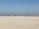 A group of people wearing colorful sports uniforms are playing cricket on a wide, open sandy ground. The sky is clear with a subtle gradient from blue to grey on the horizon. In the distance, there are structures that suggest a cityscape.