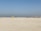 A group of people wearing colorful sports uniforms are playing cricket on a wide, open sandy ground. The sky is clear with a subtle gradient from blue to grey on the horizon. In the distance, there are structures that suggest a cityscape.