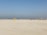 A group of people wearing colorful sports uniforms are playing cricket on a wide, open sandy ground. The sky is clear with a subtle gradient from blue to grey on the horizon. In the distance, there are structures that suggest a cityscape.