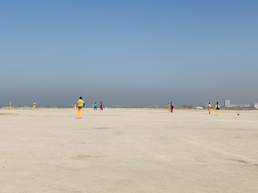 A group of people wearing colorful sports uniforms are playing cricket on a wide, open sandy ground. The sky is clear with a subtle gradient from blue to grey on the horizon. In the distance, there are structures that suggest a cityscape.
