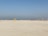 A group of people wearing colorful sports uniforms are playing cricket on a wide, open sandy ground. The sky is clear with a subtle gradient from blue to grey on the horizon. In the distance, there are structures that suggest a cityscape.