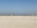A group of people wearing colorful sports uniforms are playing cricket on a wide, open sandy ground. The sky is clear with a subtle gradient from blue to grey on the horizon. In the distance, there are structures that suggest a cityscape.