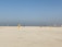 A group of people wearing colorful sports uniforms are playing cricket on a wide, open sandy ground. The sky is clear with a subtle gradient from blue to grey on the horizon. In the distance, there are structures that suggest a cityscape.