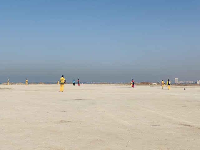 A group of people wearing colorful sports uniforms are playing cricket on a wide, open sandy ground. The sky is clear with a subtle gradient from blue to grey on the horizon. In the distance, there are structures that suggest a cityscape.