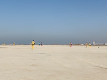 A group of people wearing colorful sports uniforms are playing cricket on a wide, open sandy ground. The sky is clear with a subtle gradient from blue to grey on the horizon. In the distance, there are structures that suggest a cityscape.