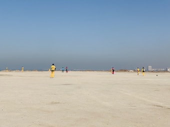 A group of people wearing colorful sports uniforms are playing cricket on a wide, open sandy ground. The sky is clear with a subtle gradient from blue to grey on the horizon. In the distance, there are structures that suggest a cityscape.
