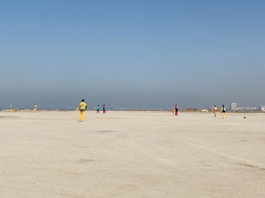 A group of people wearing colorful sports uniforms are playing cricket on a wide, open sandy ground. The sky is clear with a subtle gradient from blue to grey on the horizon. In the distance, there are structures that suggest a cityscape.