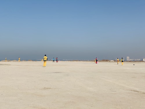A group of people wearing colorful sports uniforms are playing cricket on a wide, open sandy ground. The sky is clear with a subtle gradient from blue to grey on the horizon. In the distance, there are structures that suggest a cityscape.