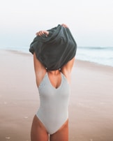 A person stands on a beach wearing a striped swimsuit, pulling a dark garment over their head. The background features a sandy shoreline and gentle waves under a pale sky.