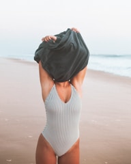 A person stands on a beach wearing a striped swimsuit, pulling a dark garment over their head. The background features a sandy shoreline and gentle waves under a pale sky.