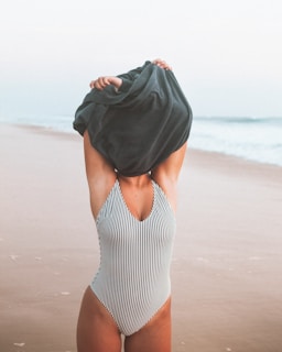 A person stands on a beach wearing a striped swimsuit, pulling a dark garment over their head. The background features a sandy shoreline and gentle waves under a pale sky.