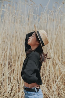 A person wearing a black top and blue jeans stands in profile against a background of tall, dry reeds. The individual is wearing a straw hat and appears to be in a relaxed pose with eyes closed and head tilted back, suggesting a sense of tranquility and enjoyment of the environment.