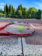 Close-up of a tennis racket and ball on a sunlit court with forest backdrop.