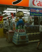 A whimsical cartoon character statue stands prominently outside a Japanese store. The scene is bustling with bright signage and vending machines filled with toys or collectibles. The interior reveals a glimpse of various products and advertisements, reflecting a vibrant commercial environment.