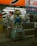 A whimsical cartoon character statue stands prominently outside a Japanese store. The scene is bustling with bright signage and vending machines filled with toys or collectibles. The interior reveals a glimpse of various products and advertisements, reflecting a vibrant commercial environment.