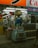 A whimsical cartoon character statue stands prominently outside a Japanese store. The scene is bustling with bright signage and vending machines filled with toys or collectibles. The interior reveals a glimpse of various products and advertisements, reflecting a vibrant commercial environment.
